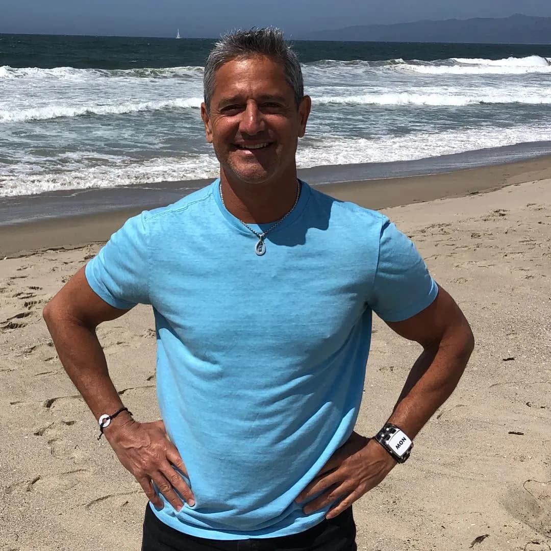 Steven on the beach in a light blue tee, hands on his hips